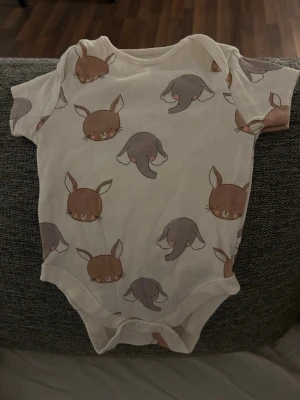 Bebis kläder Vit body med djurmönster - Supersöt vit body med korta ärmar och gulligt mönster av bruna kaniner och grå elefanter. Perfekt för dig som gillar lekfulla prints! Tryckknappar i grenen gör den enkel att ta på och av. Tillverkad i mjuk bomull för skön känsla.