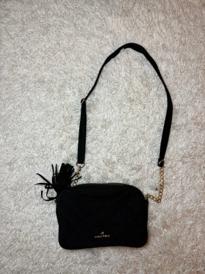 Svart quiltad axelväska från Anna Field - Säljer en svart quiltad axelväska från Anna Field med guldfärgade kedjedetaljer och dragkedja. Väskan har en mjuk mockaliknande yta, dekorativ tofs och justerbar axelrem. Perfekt storlek för det viktigaste och snygg att bära crossbody.                      Väskan är lagom använd men det finns inga tydliga tecken på slitage. 