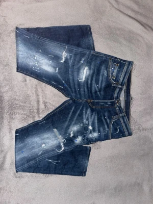 Blå jeans med slitningar från dsquared2 - Säljer ett par blå jeans med slitna detaljer och coola färgstänk. Shortsen har klassisk femficksdesign, råa kanter och en unik blå patch bak i midjan. Perfekta för dig som gillar en avslappnad och trendig stil.