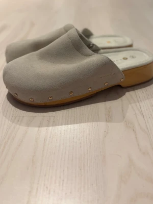 Beige tofflor i mocka från Lumia - Säljer ett par snygga beige tofflor från Lumia i mjuk mocka med träfärgad sula och guldfärgade nitar längs kanten. Perfekta att glida runt i hemma eller när du vill ha en chill look. Klassisk design med öppen häl och rund tå.