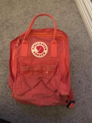 Liten kånken ryggsäck - Klassisk röd Fjällräven Kånken ryggsäck med ikonisk rund logga framtill. Ryggsäcken har ett stort huvudfack, en mindre ficka med dragkedja på framsidan och justerbara axelband. Tillverkad i slitstark syntet och har två bärhandtag upptill.                      Något urtvättad på framsidan Tillkommmer sittunderlag!