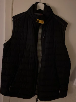 Svart dunväst från Parajumpers - Snygg svart dunväst från Parajumpers med quiltad design och hög krage. Västen har dragkedja framtill, två sidofickor med dragkedja och den klassiska gula detaljen vid halsen. Perfekt för lager på lager och håller dig varm under kyliga dagar.