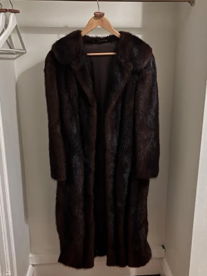 Äkta päls - Säljer en elegant lång pälsjacka i mörkbrun mink med bred krage och klassisk rak passform. Jackan har en lyxig look och är perfekt för dig som vill sticka ut med en tidlös stil. Fodrad insida för extra komfort. Superbra skick!