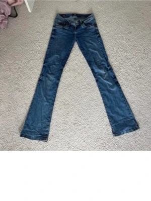 Blå bootcut jeans från LTB - Säljer ett par blå bootcut jeans från LTB med klassisk femficksmodell och snygga slitningar. Jeansen har normal midja och utställda ben, samt dekorativa sömmar på bakfickorna. Perfekta för dig som gillar en retro och avslappnad stil.
