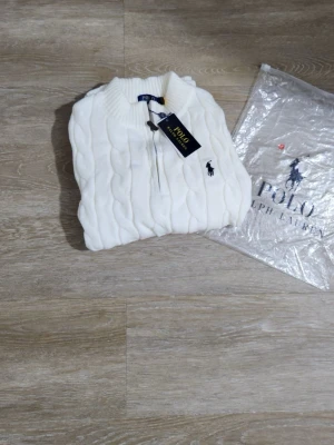 Vit kabelstickad tröja Polo Ralph Lauren - Kabelstickad vit tröja från Polo Ralph Lauren med halv dragkedja och ribbade muddar. Klassisk logga broderad på bröstet. Perfekt för dig som gillar stilrena och tidlösa plagg. Materialet känns mjukt och håller värmen.