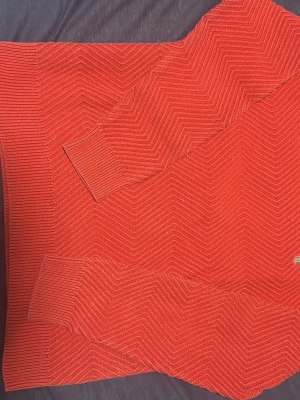 Röd stickad tröja med zigzag-mönster - Säljer en snygg röd stickad tröja med ett coolt zigzag-mönster över hela plagget. Tröjan har rund halsringning, långa ärmar och ribbade muddar vid ärmslut och nederkant. Perfekt för dig som vill ha något stilrent men ändå unikt i garderoben.