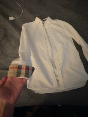 Burberry Vit skjorta med rutiga detaljer Burberry - Klassisk vit skjorta från Burberry med ikoniska beige, röda och svarta rutiga detaljer på insidan av manschetterna. Skjortan har knappar framtill, lång ärm och en stilren krage. Perfekt för dig som gillar tidlös design med en twist.