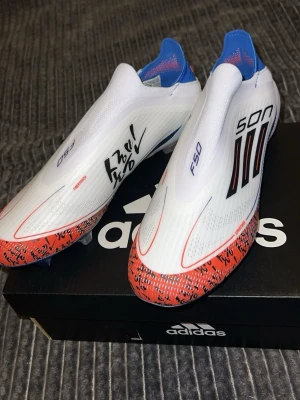 Adidas F50 SON fotbollsskor vit/röd - Säljer ett par Adidas F50 SON fotbollsskor i vitt med röda och blå detaljer. Skorna har en strömlinjeformad design utan snörning, svarta ränder på sidan och SON-tryck. Tillverkade i syntetmaterial och har autograferade detaljer på ovandelen.
