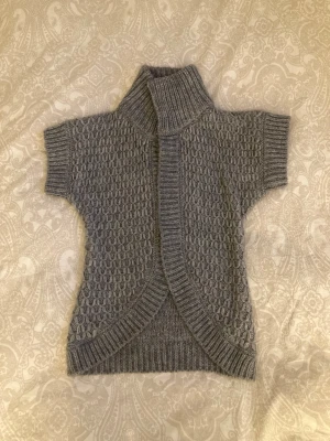 Grå stickad kofta med kort ärm - Snygg grå stickad kofta med korta ärmar och hög ribbad krage. Koftan har ett strukturerat mönster och breda ribbade kanter framtill och nedtill. Står ingen specifik storlek men skulle säga xs-m. Skriv om du vill ha fler bilder eller några mått!
