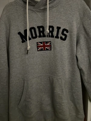 Grå hoodie från Morris med tryck - Snygg grå hoodie från Morris med svart text och brittisk flagga på bröstet. Klassisk känguruficka framtill och vita dragsnören i huvan. Tillverkad i mjukt sweatshirtmaterial, perfekt för en avslappnad stil. Det ända är att det är lite sönder vid det ena snöret