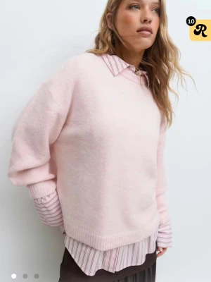 Ljusrosa stickad tröja i oversized modell - Mysig ljusrosa stickad tröja i oversized passform med rund halsringning och ribbade muddar. Tröjan har en enkel design utan mönster och är perfekt att styla lager på lager. Materialet ser ut att vara mjukt och fluffigt, troligen en ull- eller akrylblandning.