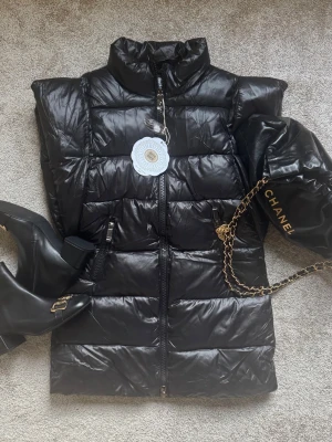 Svart dunväst  - Snygg svart dunväst med hög krage och dragkedja framtill. Västen har quiltad design och två fickor med dragkedja. Perfekt för lager på lager och ger en cool, trendig look till din outfit.