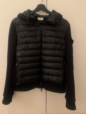 Svart dunjacka från Moncler med huva - Snygg svart dunjacka från Moncler med quiltad front, huva och dragkedja. Jackan har långa ärmar i mjukare material och en Moncler-logga på ärmen. Perfekt för kyliga dagar och ger en sportig men stilren look.