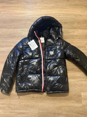 Svart pufferjacka från Moncler - Säljer en svart glansig pufferjacka från Moncler med huva och ikonisk logga på bröstet. Jackan har dragkedja med röd, vit och blå kant, flera fickor och coolt serietryck på insidan. Perfekt för dig som vill ha en varm och snygg vinterjacka.