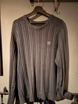 Grå kabelstickad tröja Lexington - Säljer en grå kabelstickad tröja från Lexington med rund hals och diskret logga på bröstet. Tröjan har långa ärmar och ribbade muddar vid ärmslut och nederkant.