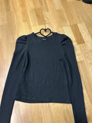 Svart långärmad topp med puffärm - Trendig svart långärmad topp från Lindex med puffiga axlar och strukturerat tyg. Rund halsringning och rak passform ger en clean look. Perfekt att styla med jeans eller kjol för en cool vardagsoutfit. Endast använd enstaka gånger.