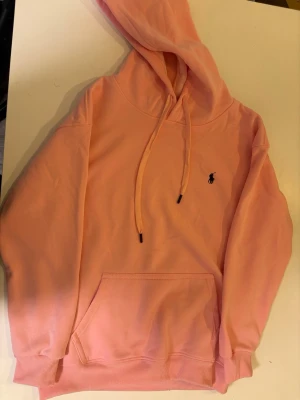 Ljusrosa hoodie från Ralph Lauren - Snygg ljusrosa hoodie från Ralph Lauren med klassisk broderad logga på bröstet. Hoodien har justerbar dragsko i huvan och en stor magficka framtill. Tillverkad i mjukt bomullsmaterial och har ribbade muddar vid ärmslut och nederkant. Liten i storlek sä rekommenderas för Small användare. Replic hoodie