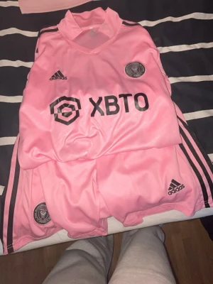 Inter Miami rosa matchställ Adidas - Säljer ett rosa matchställ från Inter Miami CF med svart logga och sponsortryck. Setet består av tröja och shorts i lätt och ventilerande material, perfekt för fotboll. Adidas klassiska ränder på axlar och sidor. Klubbmärke på både tröja och shorts.