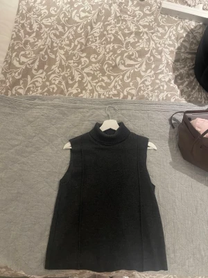 Mörkgrå stickad väst från Zara - Mörkgrå stickad väst från Zara med hög, ribbad polokrage och ärmlös design. Perfekt att bära över en skjorta eller tröja för en trendig lager-på-lager-look. Mjuk och skön kvalitet som ger en stilren och modern känsla.