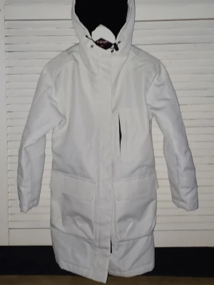 Vit parkasjacka med huva - Stilren vit parkasjacka från Lipp, med lång passform och stor huva. Jackan har dragkedja framtill, stora fickor och en bröstficka. Perfekt för kalla dagar och enkel att matcha med olika outfits. Storlek XS,  mycket stor i storleken.