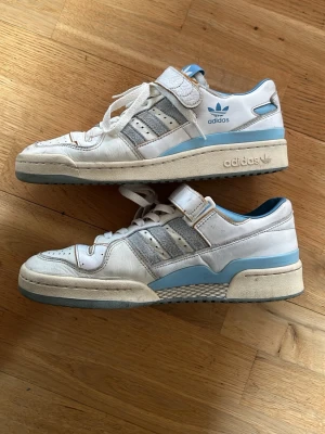 Adidas Forum Low vita/blå sneakers - Klassiska Adidas Forum Low sneakers i vitt och ljusblått med grå detaljer. Skorna har snörning, kardborreband och perforerad tå. Adidas-logga på sidan och tungan. Tillverkade i skinn och syntetmaterial med platt sula och rund tå.