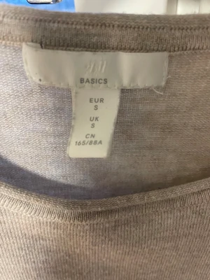 Beige stickad tröja med knappar H&M - Beige stickad tröja från H&M Basics i storlek S. Rund halsringning och korta ärmar. Snygga mörka knappar på ena axeln ger en extra detalj. Perfekt för dig som gillar enkel och stilren design med en twist.