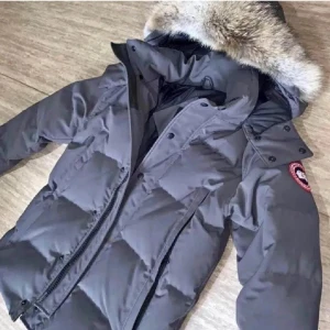 Grå Canada Goose dunjacka med päls - Säljer en grå dunjacka från Canada Goose med fet päls på huvan och klassisk röd logga på ärmen. Jackan har dragkedja, knappar och stora fickor framtill. Perfekt för kalla dagar med sin tjocka vaddering och stilrena look.