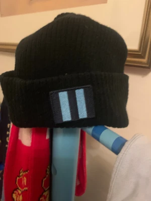 Säsongsmössa 2025 - Svart ribbstickad mössa med uppvikt kant och ett broderat märke framtill med två ljusblå ränder. Perfekt för kalla dagar och enkel att matcha med olika outfits.