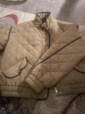 Quiltad beige jacka från Vero Moda - Quiltad beige jacka från Vero Moda med svarta kantdetaljer och guldiga knappar. Jackan har dragkedja, tryckknappar och två fickor med lock. Perfekt för dig som gillar stilren och enkel design med lite edge.