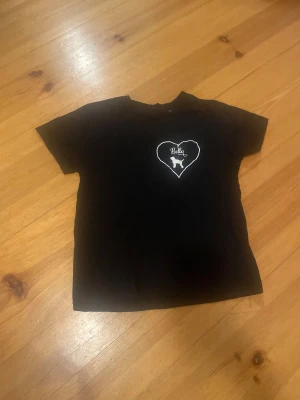 Svart t-shirt med hjärta och hund - Svart t-shirt i bomull med korta ärmar. På bröstet finns ett vitt hjärta med texten 'Bella' och en siluett av en hund. Enkel och stilren design som passar dig som gillar djurmotiv.