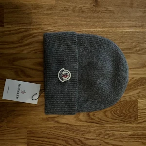 Grå ribbstickad mössa från Moncler - Snygg grå ribbstickad mössa från Moncler med klassisk uppvikt kant och broderad logotyp framtill. Tillverkad i mjuk ull som håller dig varm under kyliga dagar. Perfekt accessoar för en stilren vinterlook.