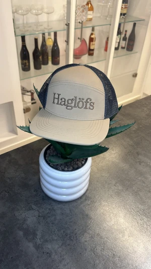 Haglöfs keps - Snygg keps från Haglöfs i beige framtill med broderad logga och marinblått mesh-tyg på sidor och bak. Justerbar snapback baktill för perfekt passform. Perfekt för dig som gillar outdoor och streetstyle.