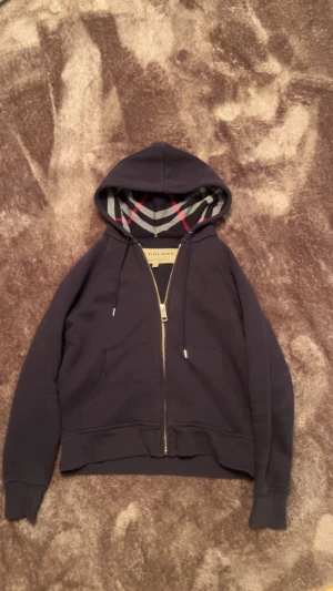 Mörkblå Burberry hoodie med dragkedja - Snygg mörkblå hoodie från Burberry med klassiskt rutigt foder i huvan och guldfärgad dragkedja. Tröjan har snörning vid huvan, två fickor fram och är långärmad. Perfekt för dig som gillar exklusiva streetwear-vibbar. Pris kan diskuteras 