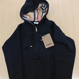 Svart hoodie från Burberry med dragkedja - Svart hoodie från Burberry med klassiskt rutigt foder i huvan och guldfärgade detaljer på snörningen. Tröjan har dragkedja framtill och en skön, loose passform. Perfekt för dig som gillar stilrena märkesplagg med ikoniska detaljer.