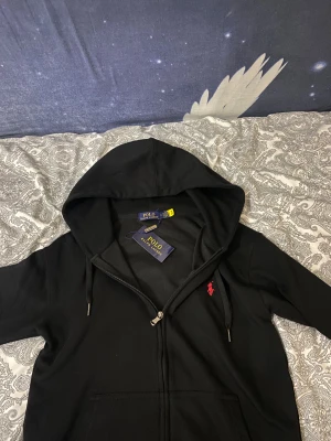 Svart hoodie från Polo Ralph Lauren - Svart hoodie från Polo Ralph Lauren med röd broderad logga på bröstet. Tröjan har huva med snörning och dragkedja framtill. Materialet är mjukt och känns som bomull, perfekt för chill dagar. Klassisk och stilren design med ficka framtill.