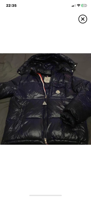 Mörkblå Moncler pufferjacka M - Säljer en mörkblå pufferjacka från Moncler i storlek M. Jackan har glansig yta, huva, dragkedja och klassisk Moncler-logga på bröstet. Insidan har en cool serietidningsdetalj och röd-vit-blå rand vid dragkedjan. Perfekt för kalla dagar.