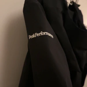 Svart vindjacka Peak Performance - Svart vindjacka från Peak Performance med huva och hel dragkedja framtill. Jackan har två sidofickor med dragkedja och diskret logga på ärmen. Materialet är lätt och syntetiskt, perfekt för blåsiga dagar. Stilren och sportig design som funkar till många outfits.