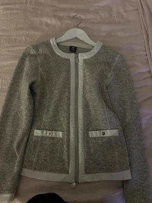 Grå cardigan jacka med dragkedja - Snygg grå cardigan jacka från KappAhl med ljusa detaljer och dragkedja framtill. Jackan har rund hals, två fickor med knapp och ljus kant vid fickor, hals och nederkant. Perfekt för dig som gillar stilren och enkel design.