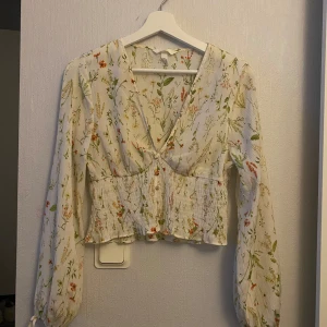 Blommig vit blus med v-ringning - Superfin blommig blus i vit med v-ringning och smockad midja. Långa ärmar med lätt puff och knyt vid ärmslut. Blusen har små tygklädda knappar framtill och ett romantiskt blommönster i grönt, orange och rosa. Perfekt för vår och sommar!