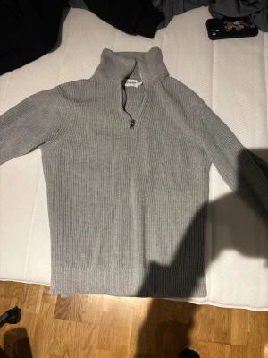 Grå ribbad half zip stickad tröja - Snygg grå stickad tröja från East West med ribbad struktur och half zip i halsen. Perfekt för dig som gillar en clean look med lite extra edge. Hög krage och långa ärmar gör den både stilren och chill. Passar dig som vill ha något enkelt men ändå trendigt.