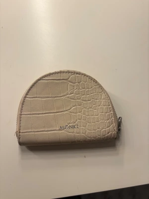 Beige plånbok med croco-mönster från Monki - Snygg beige plånbok från Monki i halvmåneformad design med croco-mönstrad yta. Plånboken har dragkedja i silver och flera fack inuti för kort och småsaker. Perfekt att ha med sig i väskan eller fickan för en clean och trendig look.
