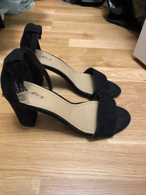Svarta klackade sandaler från Alley - Svarta sandaler från Alley med bred rem över tårna och justerbart spänne runt ankeln. Skorna har en stadig blockklack och öppen tå. Utsidan är i mockaliknande material och innersulan är beige. Perfekta för att lyfta din outfit.