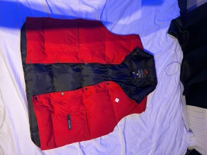 Röd dunväst från Canada Goose XXL - Snygg röd dunväst från Canada Goose med svart foder och klassisk logga med lönnlöv på bröstet. Västen har praktiska fickor framtill, dragkedja och tryckknappar. Perfekt för kalla dagar när du vill ha både stil och värme.
