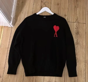 Svart stickad tröja från Ami - Svart stickad tröja från Ami Alexandre Mattiussi med röd hjärtlogga och bokstaven A på bröstet. Tröjan har rund hals, ribbade muddar och långärmad passform. Perfekt för dig som gillar stilrena plagg med en twist.