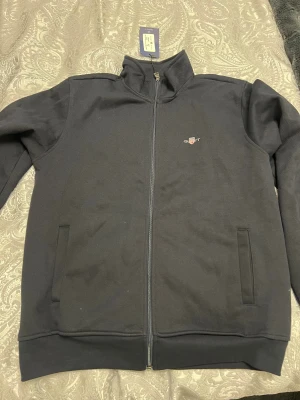 Svart ziptröja från Gant - Snygg svart ziptröja från Gant med broderad logga på bröstet. Tröjan har hög krage, hel dragkedja och två fickor framtill. Perfekt för dig som gillar stilrena och klassiska plagg. Materialet är mjukt och känns skönt mot huden. Forfarandd etikett finns