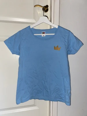 Blå t-shirt med guldkrona  - Snygg ljusblå t-shirt från Fruit of the Loom med en glittrig guldkrona tryckt på bröstet. 👑Materialet är mjuk bomull som känns skönt mot huden. Den har aldrig kommit till användning, storlek L men liten i storleken 👑 