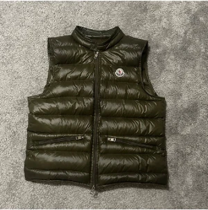 Olivgrön dunväst från Moncler - Snygg olivgrön dunväst från Moncler med quiltad design och hög krage. Västen har två fickor med dragkedja framtill och klassisk Moncler-logga på bröstet. Perfekt att slänga över en hoodie för extra värme. Materialet är glansigt och känns lyxigt. PRIS KAN DISKUTERAS!!