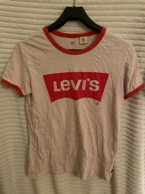 Vit och röd Levi's t-shirt - Säljer en klassisk vit t-shirt från Levi's med rött tryck och logga på bröstet. T-shirten har röda detaljer vid krage och ärmslut, samt korta ärmar. I mycket bra skick och säljs i befintlig skick. 