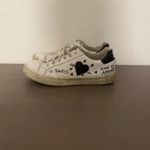 Vita sneakers med svart hjärtdetalj - Unika vita sneakers med svart häl och handritade detaljer som hjärta, stjärnor och texten 'Paris Mon Amour' på. Perfekta för dig som vill sticka ut! Har ni någon fråga är det bara att fråga på! 
