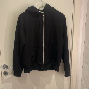 Svart hoodie med dragkedja från H&M - Svart hoodie från H&M i storlek M med huva och snörning. Tröjan har en praktisk dragkedja framtill och två stora fickor. Perfekt för chill dagar och enkel att matcha med jeans eller joggers.